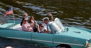 Guests cruise across the water in a turquoise amphibious car, waving proudly with a US flag at Disney Springs, Walt Disney World.