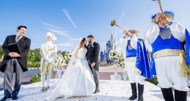 Newlyweds share a kiss in front of Cinderella Castle at Disney World, with trumpeters and an officiant creating fairytale magic.