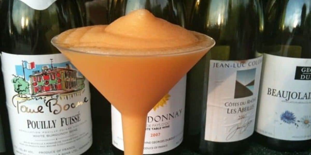 A slushy cocktail served in a martini glass, standing in front of a row of assorted wine bottles with colorful labels. The drink is orange and has a frothy texture.