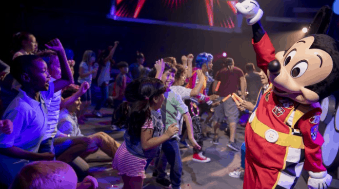 Kids dance and cheer around Mickey Mouse at the Disney Junior Dance Party, surrounded by vibrant, colorful lights indoors.