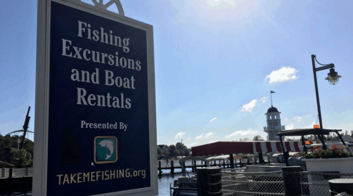Fishing Excursions and Boat Rentals sign on the marina dock at our waterfront attraction, with boats and a lakefront lodge behind.