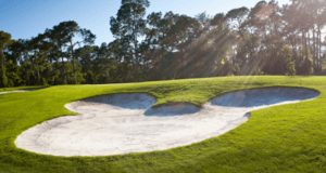 A Mickey Mouse-shaped sand bunker on a sunlit Disney World golf course, surrounded by lush green grass and trees.