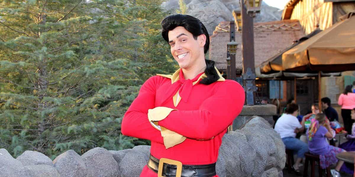 A person in a red costume with a yellow collar and black belt poses confidently with arms crossed. The background features greenery and rustic buildings with people seated nearby.
