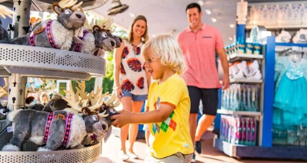 A young child in a yellow shirt excitedly reaches for a plush reindeer in a store. Two adults smile in the background. The store is filled with stuffed toys and costumes, creating a colorful and cheerful atmosphere.
