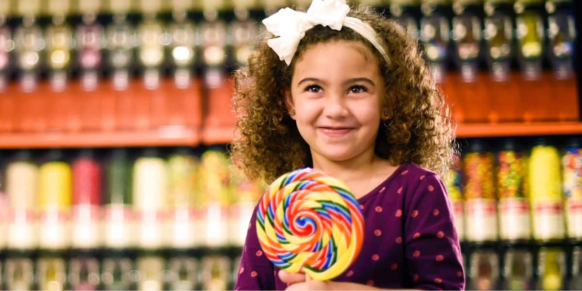 A smiling young girl with curly hair and a white bow is holding a large, colorful lollipop. She is wearing a purple polka-dot shirt and standing in front of a wall of candy jars.