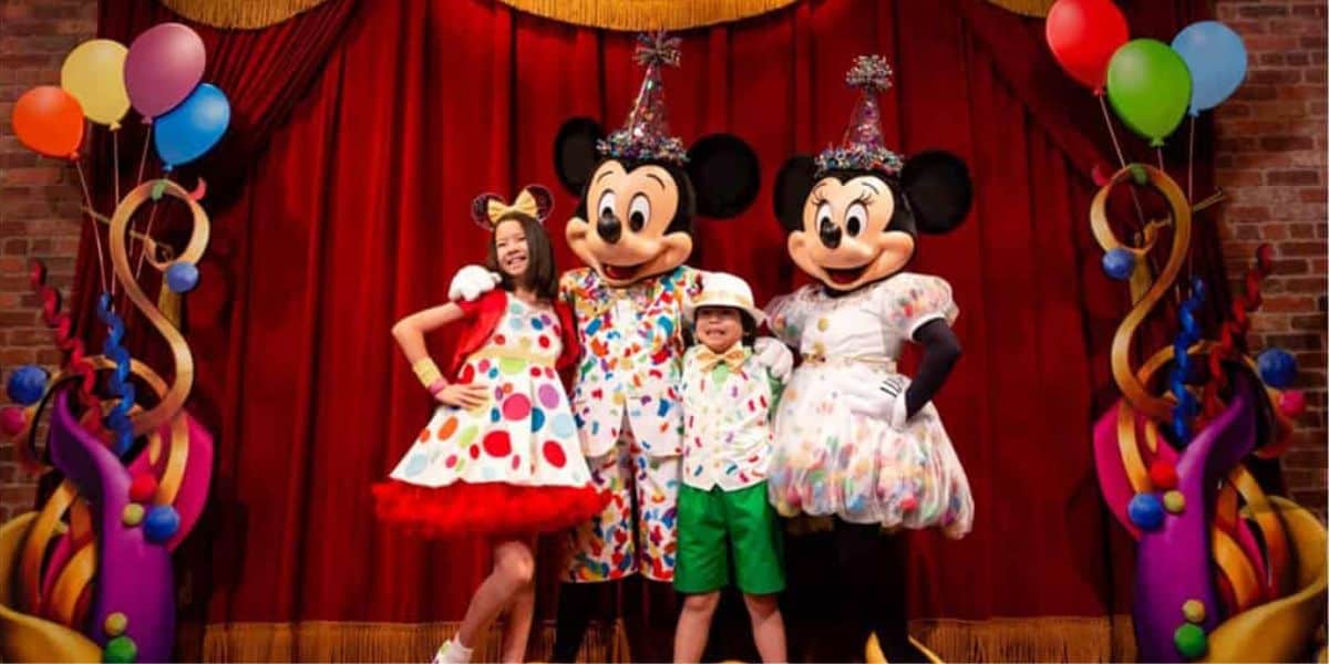 Two children in colorful party outfits pose with characters wearing festive costumes in front of a red curtain. The backdrop is decorated with colorful balloons and streamers, creating a joyful celebration atmosphere.