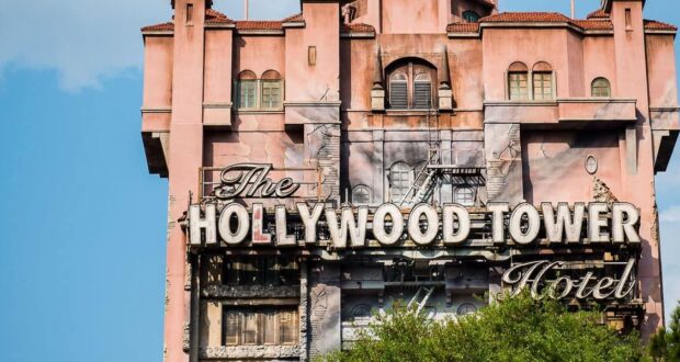 The image shows the Hollywood Tower Hotel, a tall, intricately designed building with a weathered facade and an ornate sign. The structure has a pinkish hue, and the sky is blue with a few clouds in the background. Trees partially obscure the view.