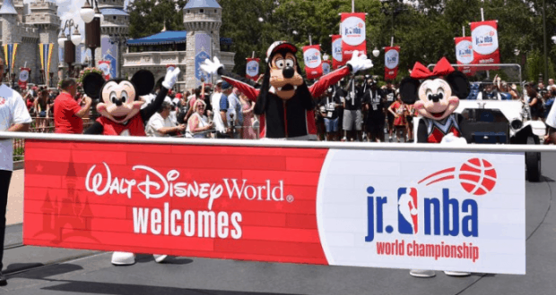 Mickey, Goofy, and Minnie celebrate at Disney World, standing behind a Jr. NBA World Championship banner.