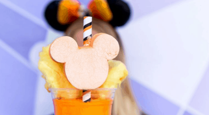Vibrant orange drink with Mickey-shaped apple slice, pineapple, and striped straw; Mickey ears in the background at Disney World.