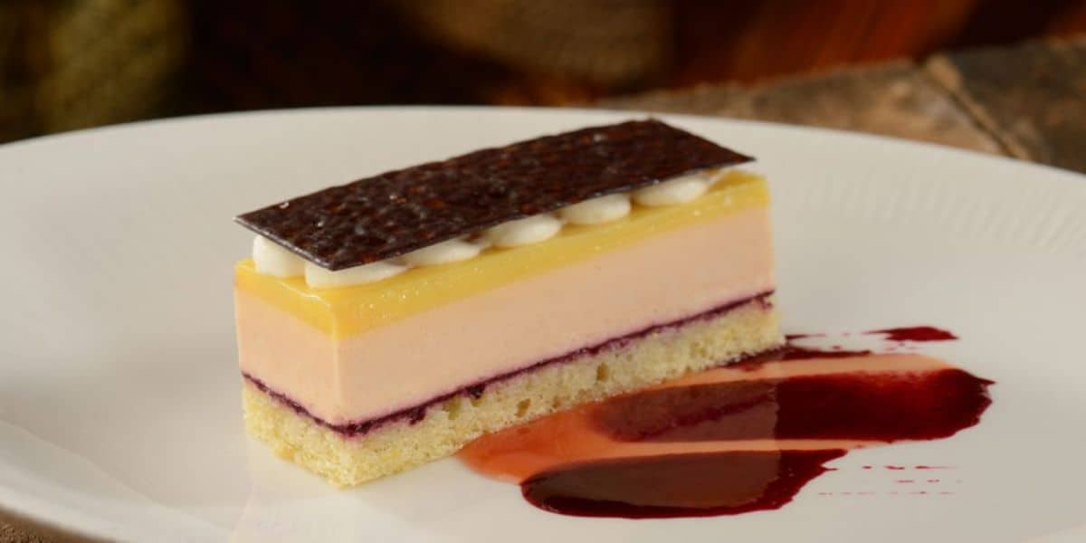 A slice of layered dessert on a white plate, featuring a sponge base, berry filling, creamy layers, and topped with a glossy chocolate sheet and dollops of cream. A berry sauce is artfully drizzled beside it.
