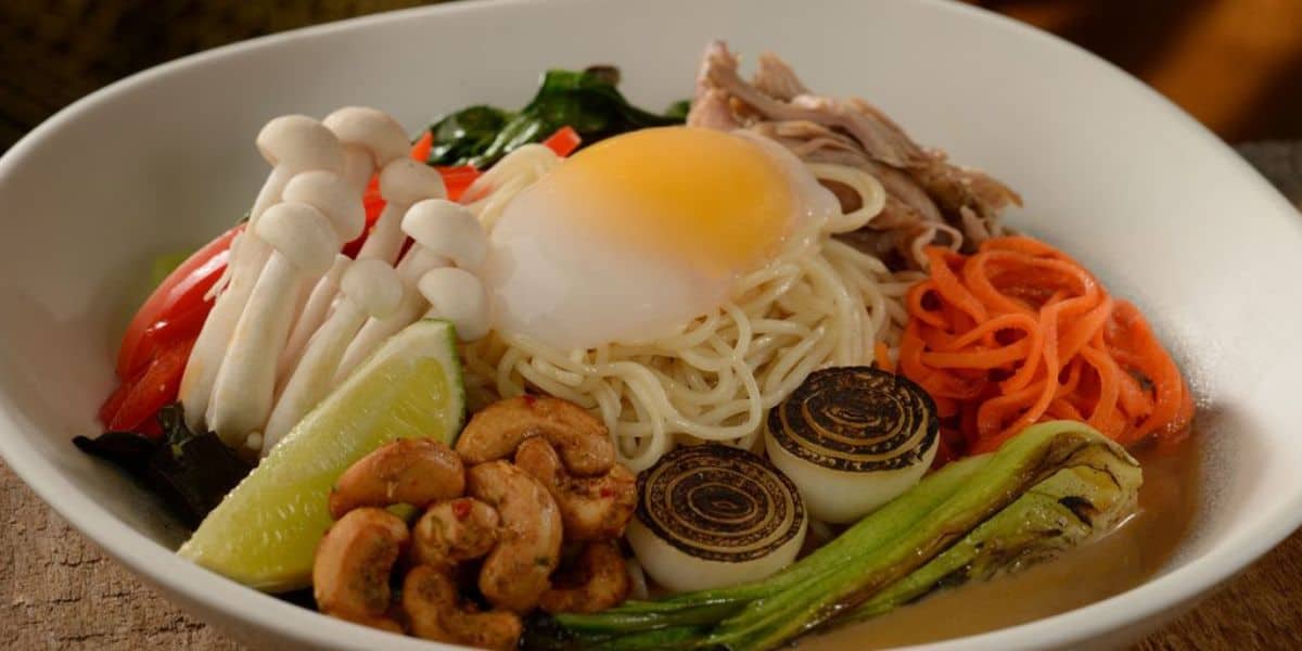A bowl of ramen with a soft-boiled egg, enoki mushrooms, sliced red bell pepper, shredded meat, carrot spirals, grilled green onions, lime wedge, cashews, radishes, and greens in broth.