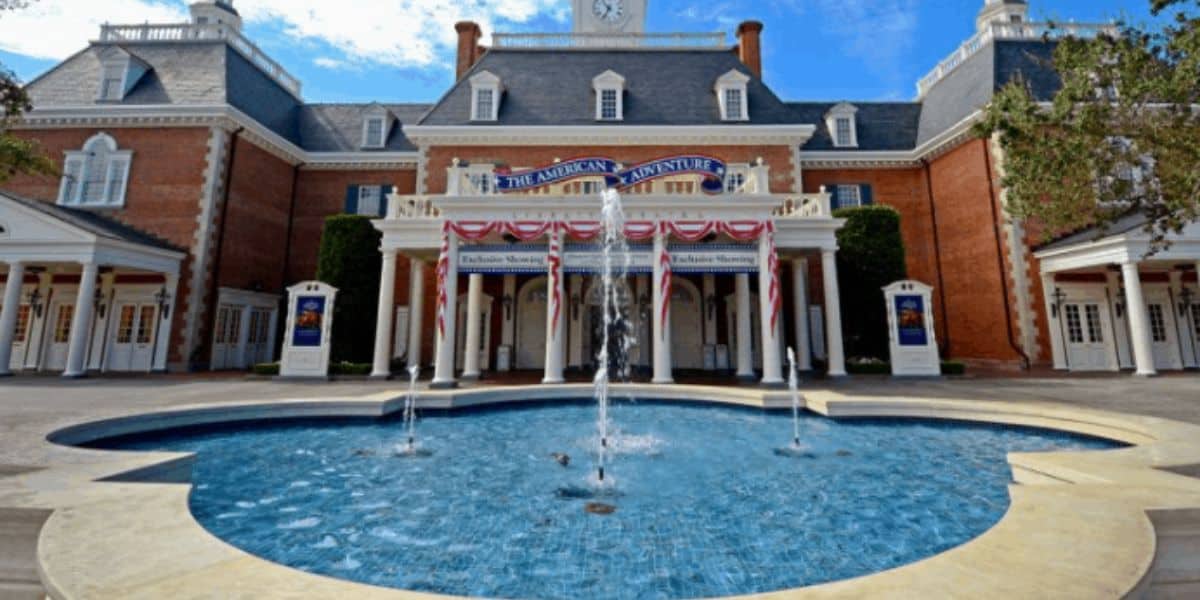 A stately brick building with a clock tower, featuring a large central fountain in front. The facade is decorated with American flags and banners, set against a clear blue sky.