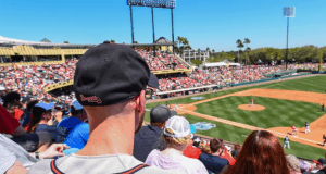 Fans enjoy Atlanta Braves Spring Training under the sun, viewed from behind a guest in a black cap at the ballpark.