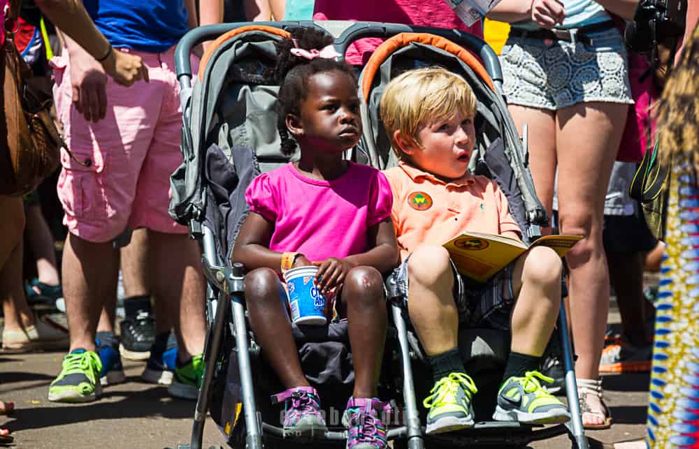 Two kids in a stroller enjoy Disney World’s excitement—one sipping a drink, the other reading, surrounded by energetic crowds.
