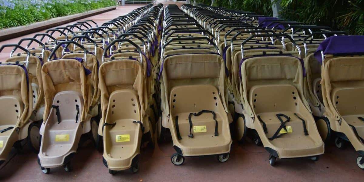 A large group of identical beige strollers neatly arranged in multiple rows on a paved pathway, surrounded by greenery. The strollers have black handles and wheels.