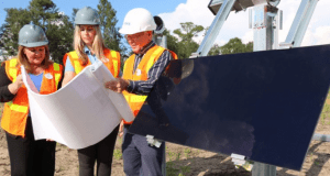 Cast Members in safety gear go over blueprints beside a solar panel at the Walt Disney World solar facility under bright sunshine.