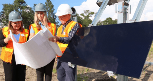 Cast Members in safety gear go over blueprints beside a solar panel at the Walt Disney World solar facility under bright sunshine.