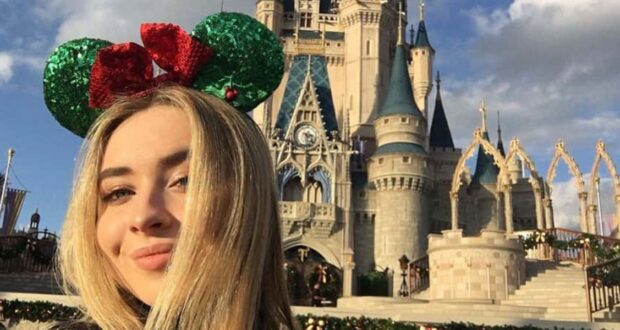 A woman with long blonde hair wears green and red Mickey Mouse ears. She is smiling in front of a castle at a theme park, with blue sky and clouds in the background.