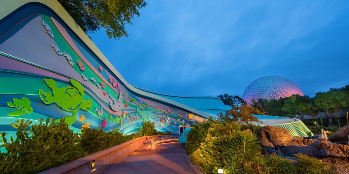 A vibrant, curved building with colorful sea-themed murals is lit up at night. In the background, a geodesic dome structure glows softly. Lush greenery surrounds the path leading to the entrance, creating a lively atmosphere.