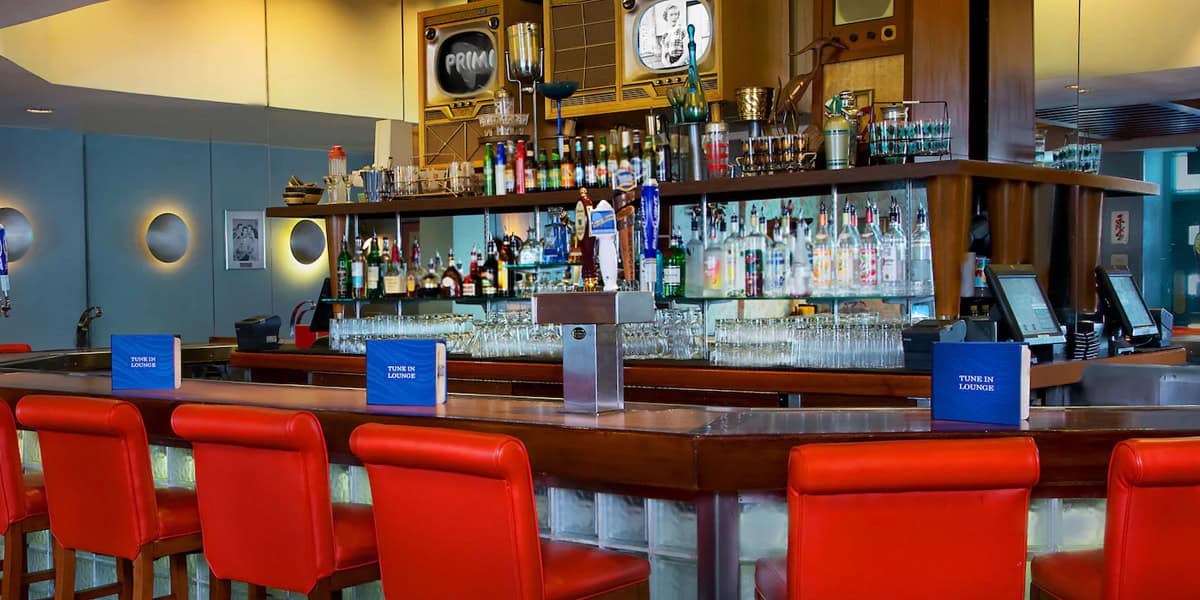 A modern bar with red barstools at a wooden counter, shelves of glassware and bottles showcasing the best alcoholic drinks at Hollywood Studios, blue menus on the counter, vintage decor, and round wall lights.