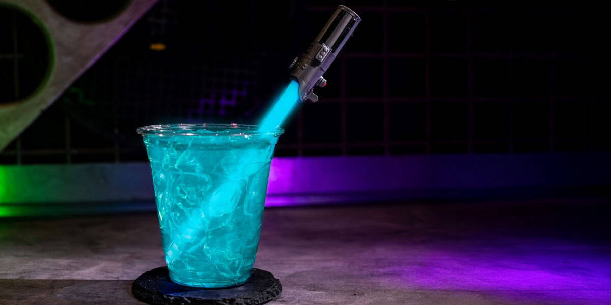 A glowing blue drink, inspired by the best alcoholic drinks at Hollywood Studios, sits in a plastic cup with a lightsaber-shaped stirring stick on a dark surface, illuminated by colorful lighting in the background.