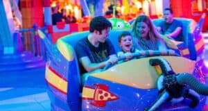 A family of three enjoys a colorful amusement park ride. The child sits between two adults, all smiling and having fun. The ride is vibrant with playful designs, and the background is filled with bright lights and other attractions.