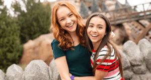 Two friends embrace by a stone fence at Disney’s Hollywood Studios, channeling Kim Possible with trees and classic park scenery behind.