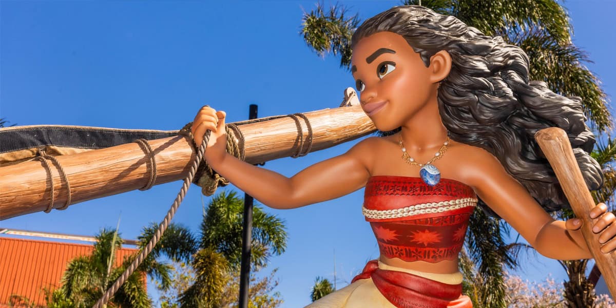 A statue of Moana, Disney character with long curly hair, wearing a red and white top and holding a wooden oar, stands outdoors at Disney’s Polynesian Village Resort with palm trees and a clear blue sky in the background.