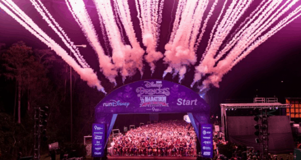 Fireworks burst above Walt Disney World's Princess Half Marathon as runners gather beneath a glowing purple start arch.