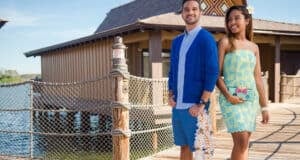 A man and woman stand on a wooden walkway by the water at Disney’s Polynesian Village Resort, near a thatched-roof building. The man wears a blue cardigan, blue shorts, and holds a lei. The woman wears a light blue and yellow dress with a small clutch.