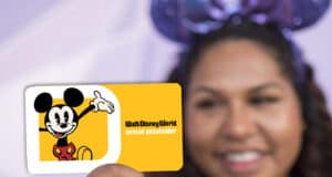 A person holds a Disney World Annual Pass card featuring Mickey Mouse on a yellow background. The person is smiling and wearing Mickey Mouse ears, ready to enjoy all the magic of being an Annual Passholder.