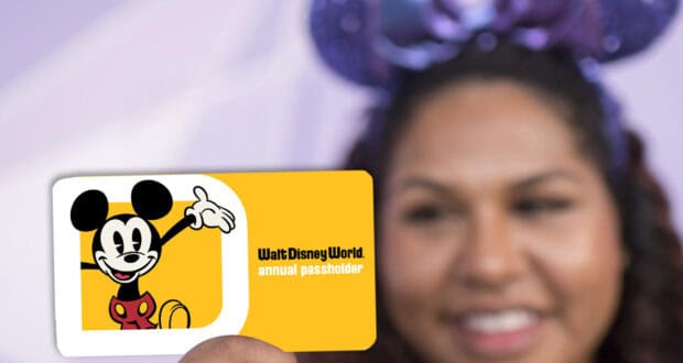 A person holds a Disney World Annual Pass card featuring Mickey Mouse on a yellow background. The person is smiling and wearing Mickey Mouse ears, ready to enjoy all the magic of being an Annual Passholder.