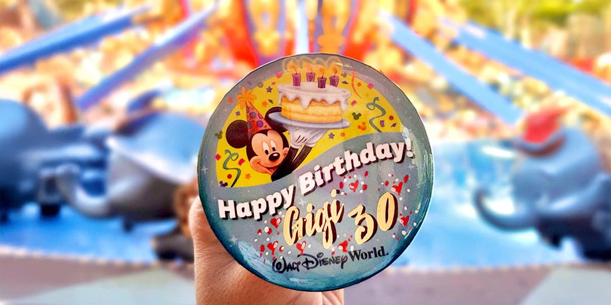 A hand holds a round Disney birthday button—one of the free things at Walt Disney World—featuring Mickey Mouse, a cake, and the words "Happy Birthday! Greg 30 Walt Disney World" with colorful decorations. Blurry theme park rides are visible in the background.