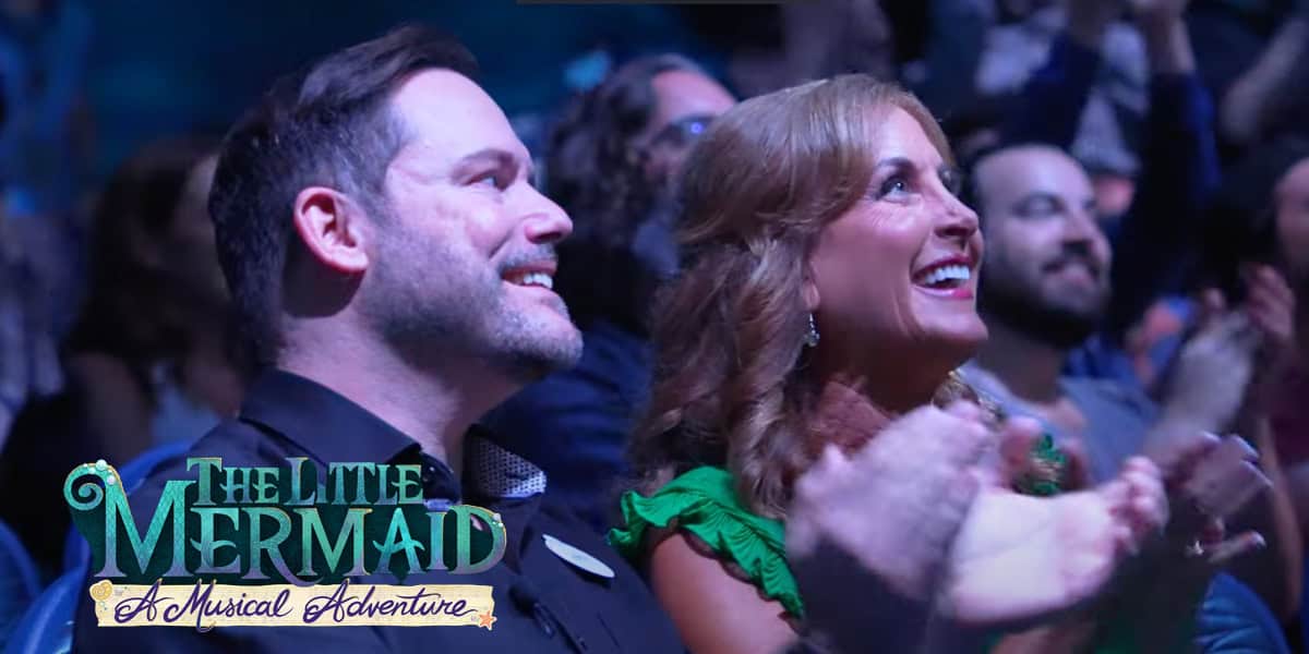Two people smile and clap enthusiastically in an audience, likely Disney World Annual Pass holders. The image is overlaid with the text "The Little Mermaid: A Musical Adventure." Other audience members are visible in the background.