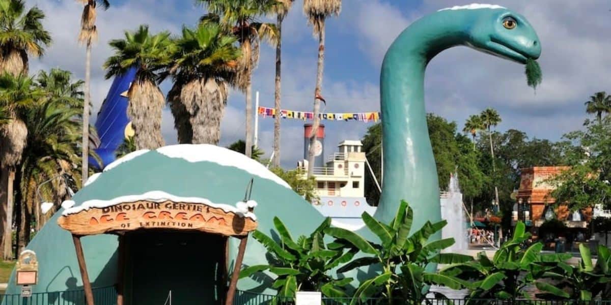 A green dinosaur statue, with a long neck and a smiling face, stands next to a dome-shaped building. The building's sign reads "Dinosaur Gertie's Ice Cream of Extinction," surrounded by palm trees and a clear blue sky.