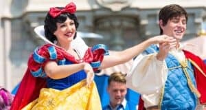 Two performers dressed as Snow White and a prince are dancing happily on a castle stage. Snow White wears a classic red, blue, and yellow dress with a red bow, while the prince wears a blue vest over a white shirt. A crowd watches in the background.
