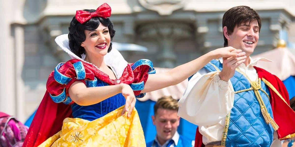 Two performers dressed as Snow White and a prince are dancing happily on a castle stage. Snow White wears a classic red, blue, and yellow dress with a red bow, while the prince wears a blue vest over a white shirt. A crowd watches in the background.