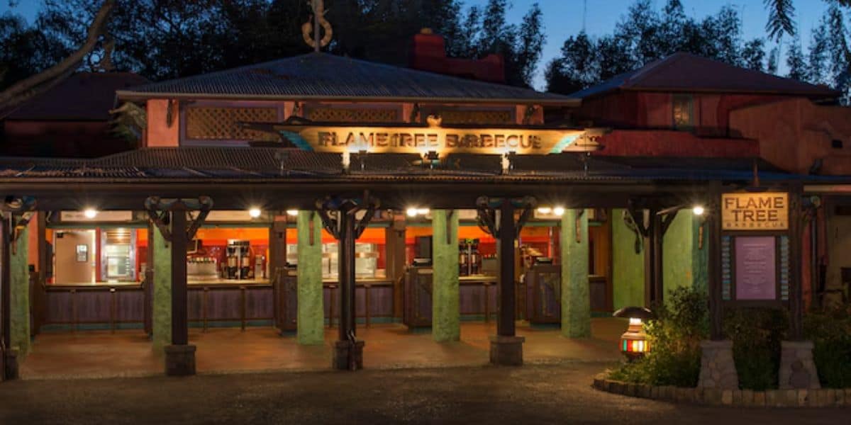 A rustic barbecue restaurant with a wooden exterior and a corrugated metal roof, warmly lit at night. The sign reads "Flame Tree Barbecue," and the outdoor area is surrounded by trees and decorative elements.