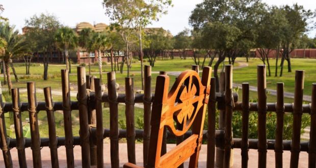 A wooden chair with a carved backrest overlooks a wide grassy field, enclosed by a rustic bamboo fence. Trees are scattered across the field, and buildings resembling Magical Disney Moments are visible in the distance under a clear sky.