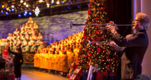 A trumpeter performs on stage beside a festive Christmas tree and robed choir during Epcot's Candlelight Processional.