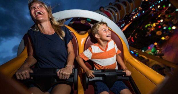 A woman and a young boy are seated on a roller coaster, both smiling and laughing with excitement. The ride is illuminated with colorful lights in the background, contrasting against a dark, cloudy sky.