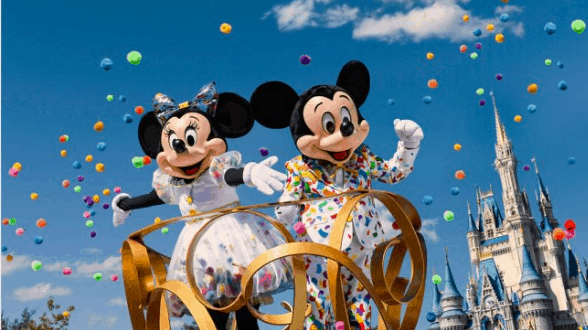 Mickey and Minnie Mouse in vibrant costumes wave on a confetti-filled parade float by Cinderella Castle at Walt Disney World.