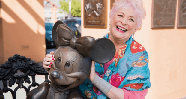 Guest with vibrant pink hair and colorful scarf poses by the Minnie Mouse statue, celebrating Russi Taylor at Disneyland.