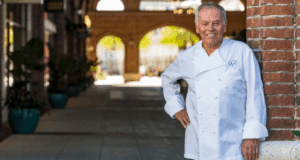 Wolfgang Puck smiles in his signature white chef coat, relaxing by a brick wall at the lively Bar & Grill on the promenade.