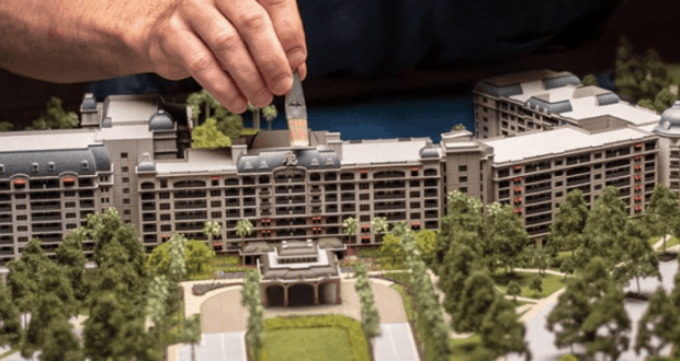 A hand adds a piece to the intricate Disney's Riviera Resort model, showcasing the stunning hotel and its lush, tree-filled grounds.