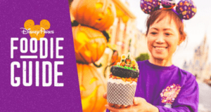 Disney fan in purple Minnie ears shows off a Halloween cupcake; Disney Parks Foodie Guide logo and pumpkins tease party treats.