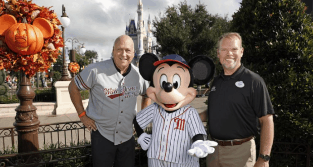 Mickey Mouse and two men in baseball gear celebrate the Ripken Experience at Disney World, with fall decor by Cinderella Castle.