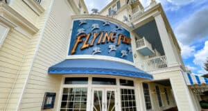 Entrance of "Flying Fish" restaurant with a nautical theme, featuring a blue awning and decorative fish accents. The building is cream-colored with large windows and balconies. The sky above is partly cloudy.