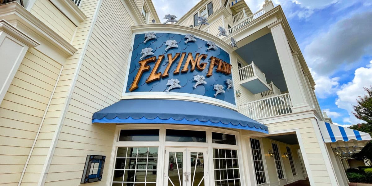 Entrance of "Flying Fish" restaurant with a nautical theme, featuring a blue awning and decorative fish accents. The building is cream-colored with large windows and balconies. The sky above is partly cloudy.