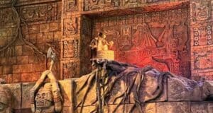 A detailed ancient temple wall with intricate carvings and symbols. A sculpture of a figure holding a staff stands atop a rock formation. The scene is lit with warm, dramatic lighting, highlighting the carvings and textures.